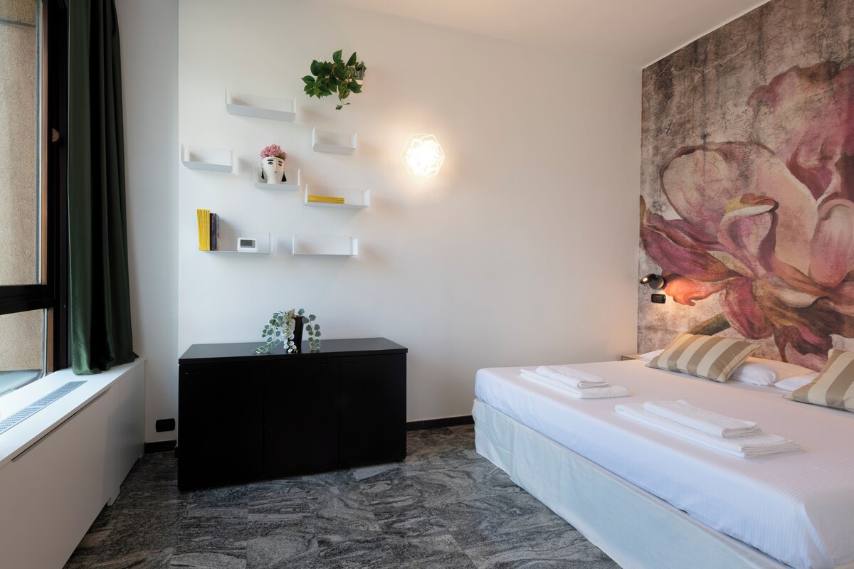 Quarto de hotel com uma cama, um armário preto, uma estante fixada na parede e um quadro floral grande.