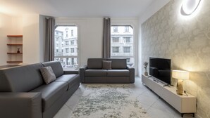 Een moderne woonkamer met een bank, een kleinere zithoek, een televisie en een lamp op een bijzettafeltje.