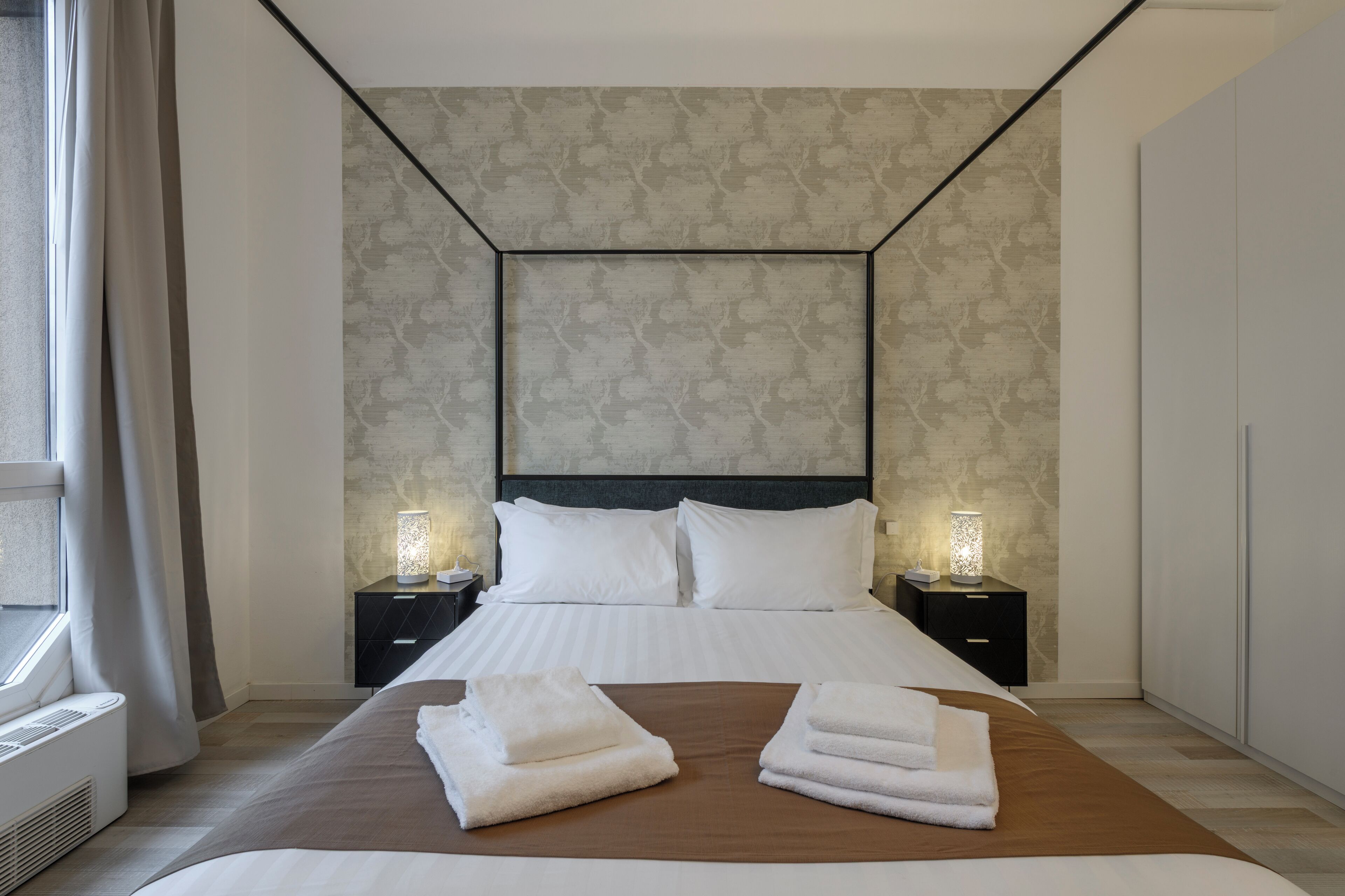 A modern hotel room with a large bed, two bedside tables with lamps, a wardrobe, and a window with curtains.