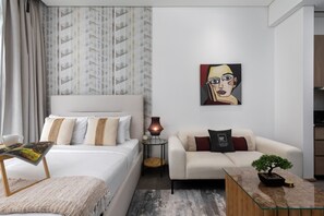 Quarto de hotel moderno com cama, sofá, mesa de centro com um bonsai, abajur e um quadro na parede.