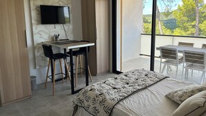 A modern bedroom with a bed, a desk, and chairs. There is a TV on the wall and a balcony with a view of trees.