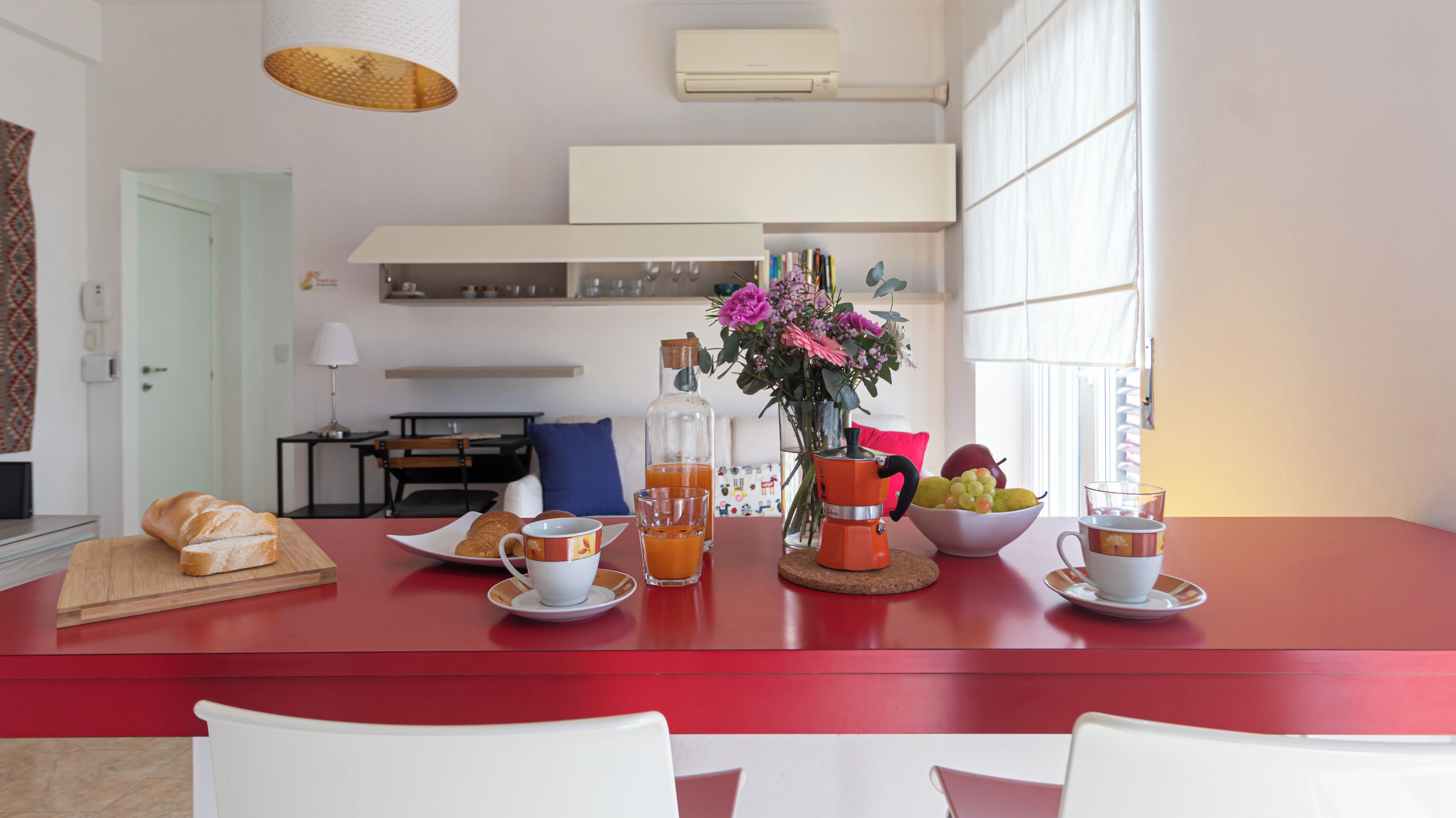 A dining area with a red table, white chairs, a bottle of orange juice, a bowl of fruit, and a vase with flowers.