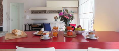 A dining area with a red table, white chairs, a bottle of orange juice, a bowl of fruit, and a vase with flowers.