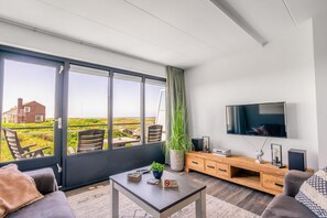 A modern living room with a large window, a flat-screen TV, a wooden entertainment unit, and a potted plant. - 4-person Apartment With Island-sea View (De Koog)