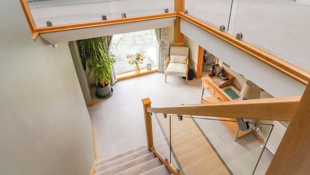 Un intérieur moderne avec un escalier menant à un niveau inférieur. On y trouve une plante en pot, une chaise et une petite table avec des livres et une lampe.