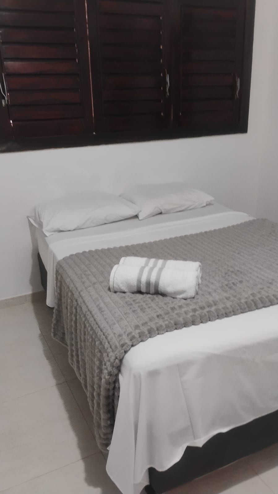 A bed with a grey textured bedspread and white pillows, a white blanket, and a rolled white towel on the bed.