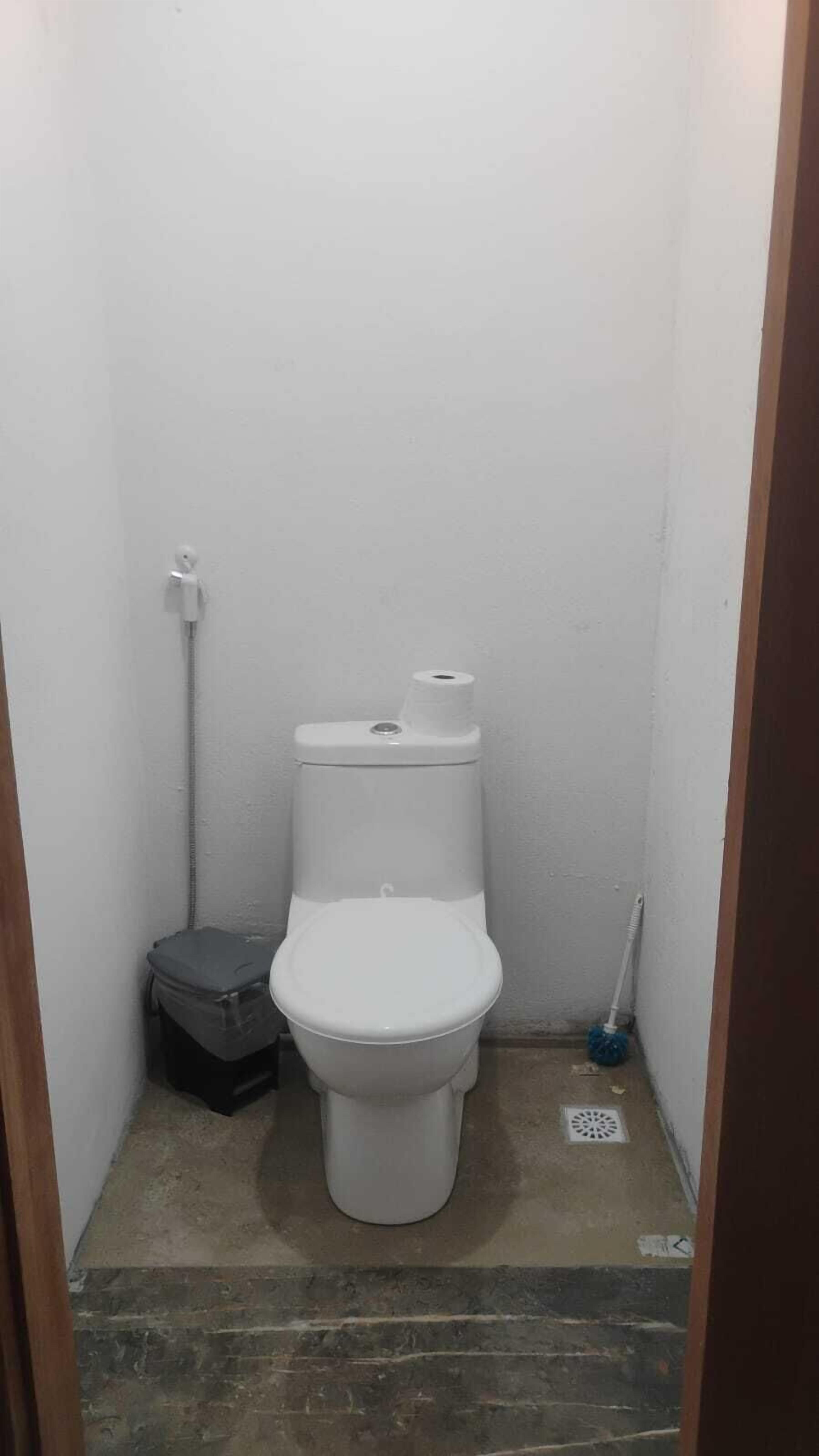 Un bany petit i estret amb un vàter blanc, un paperer i una escoba.