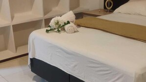 Een net opgemaakt bed met witte lakens en een klein boeket witte bloemen, een nachtkastje met een lamp, en een witte wandplank.