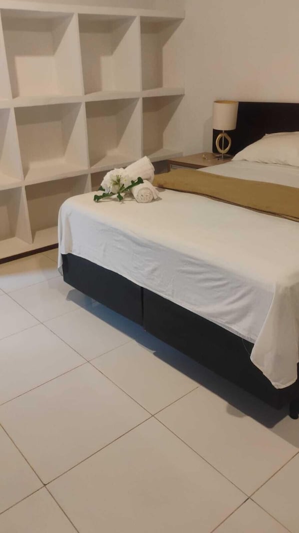 A neatly made bed with white linens and a small bouquet of white flowers, a bedside table with a lamp, and a white shelving unit against the wall. - Pousada Sol de Maria (Tibau do Sul)