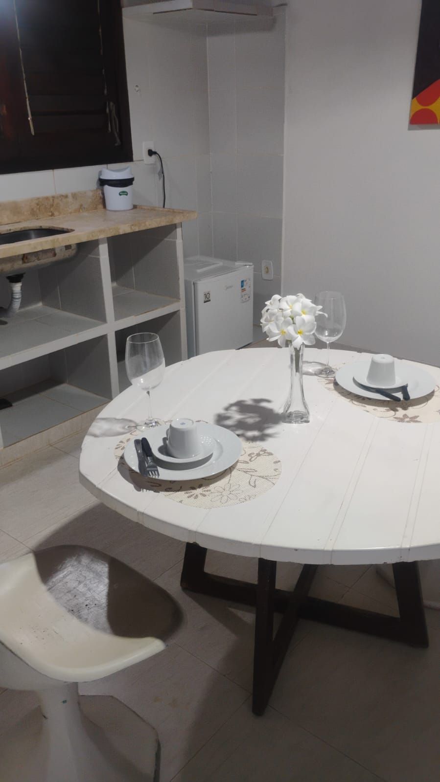 A round dining table with a white tablecloth, two white plates, a vase with white flowers, and two wine glasses.