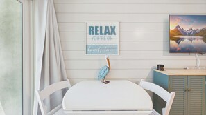 A room with a white table, a framed sign saying 'RELAX YOU'RE ON beach time', a pelican decoration, a painting of mountains, and a cabinet with a book.