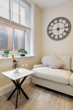 A room with a white sofa, a round wall clock, a window with blinds, a small table with a champagne bottle and glasses, and two potted plants on the windowsill. - Tottenham Court's Road - 2Bed Apartment (London)