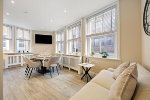 A modern living room with a beige sofa, a dining area with a round table and chairs, large windows with blinds, and a mounted TV. - Tottenham Court's Road - 2Bed Apartment (London)