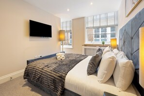 A modern bedroom with a large bed, a flat-screen TV mounted on the wall, and a window with blinds. - Tottenham Court's Road - 2Bed Apartment (London)