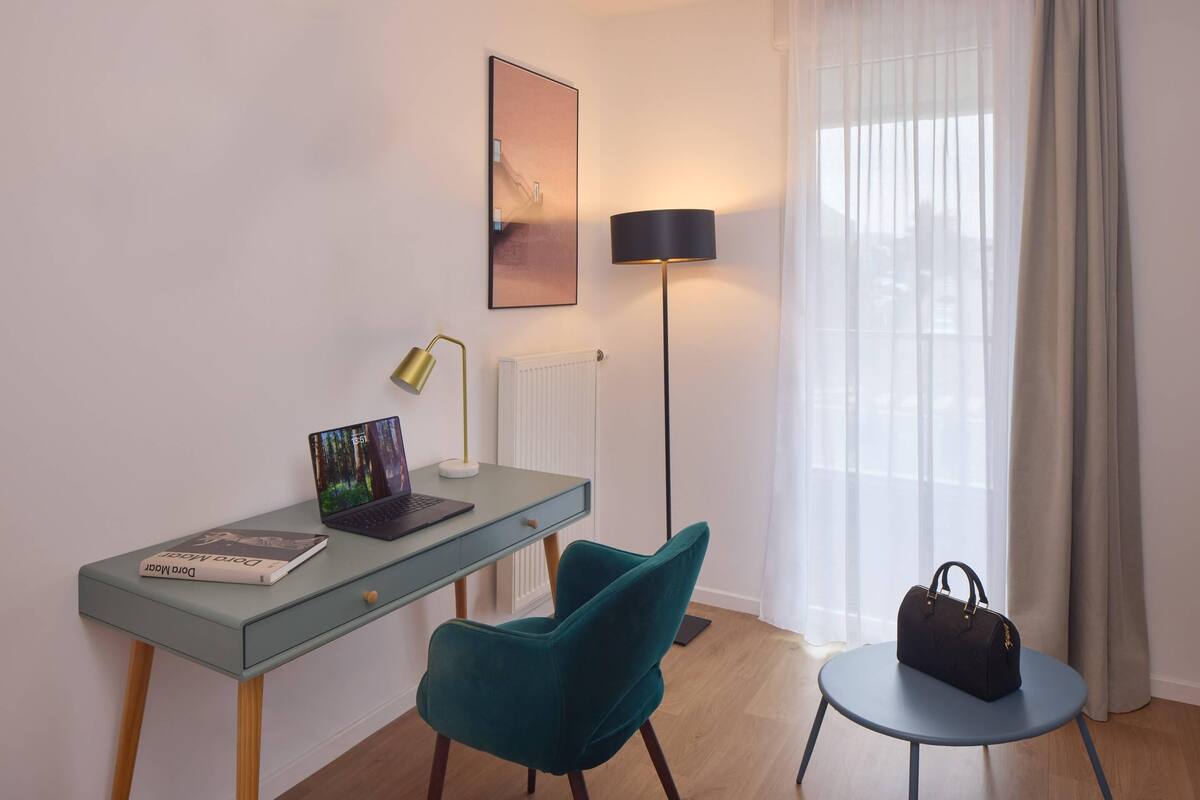 A modern room with a glass desk, a teal chair, a round table, a lamp, a radiator, and a window with curtains.