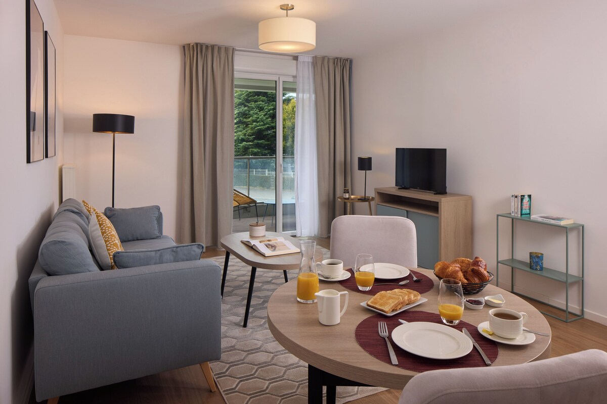 A modern hotel room with a sofa, dining table, breakfast setup, TV, and a balcony with curtains.