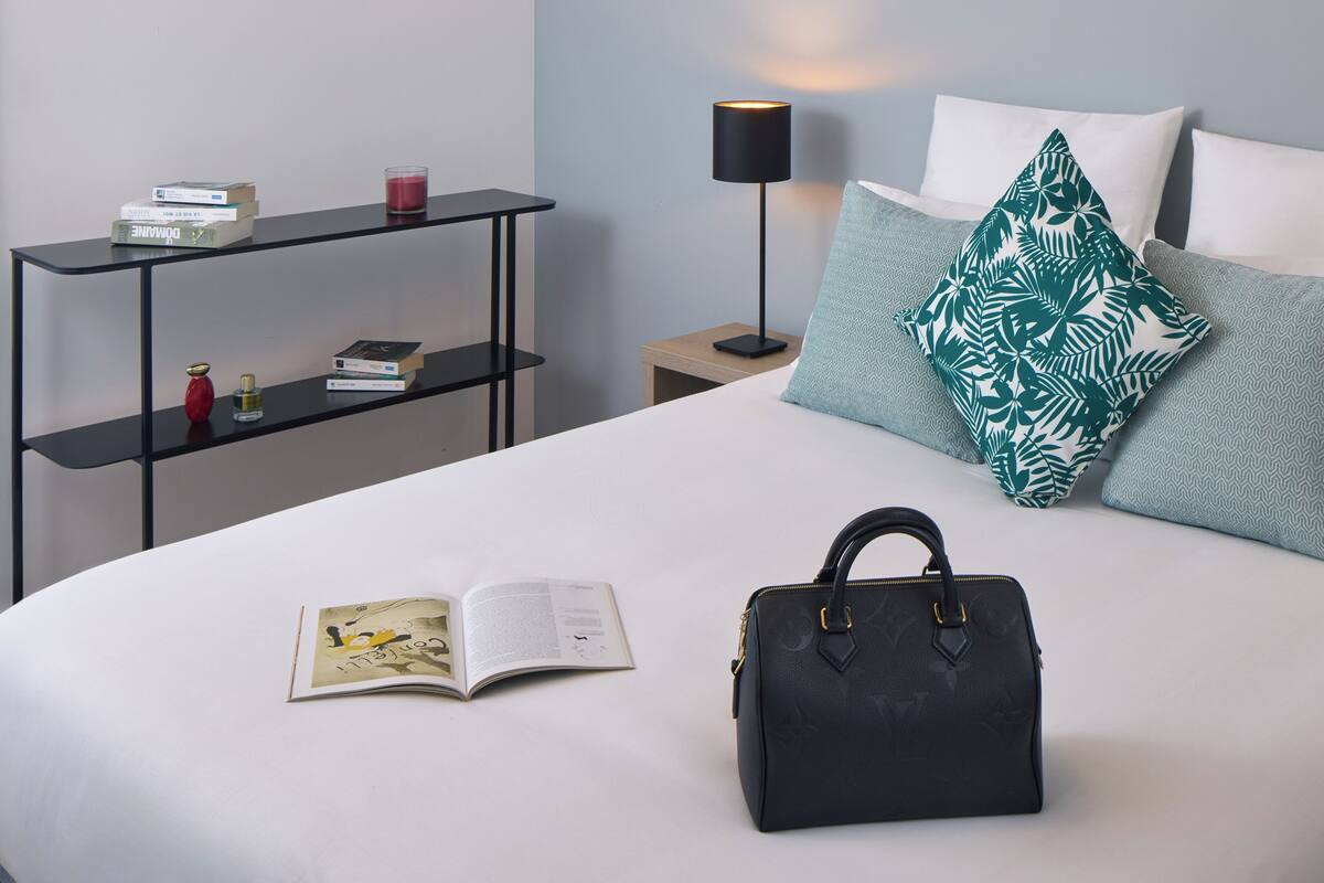 A neatly made bed with a book, a handbag, a nightstand with a lamp, and a shelf with books and a candle.