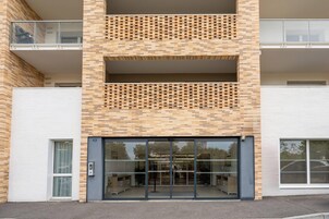 Un edificio moderno con fachada de ladrillo, grandes puertas de cristal de entrada y un balcón visible en el piso superior.