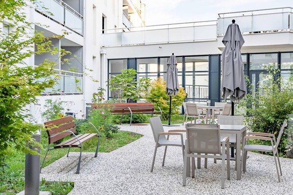 Un espacio al aire libre con mesas y sillas, rodeado de follaje y un edificio con ventanas amplias.