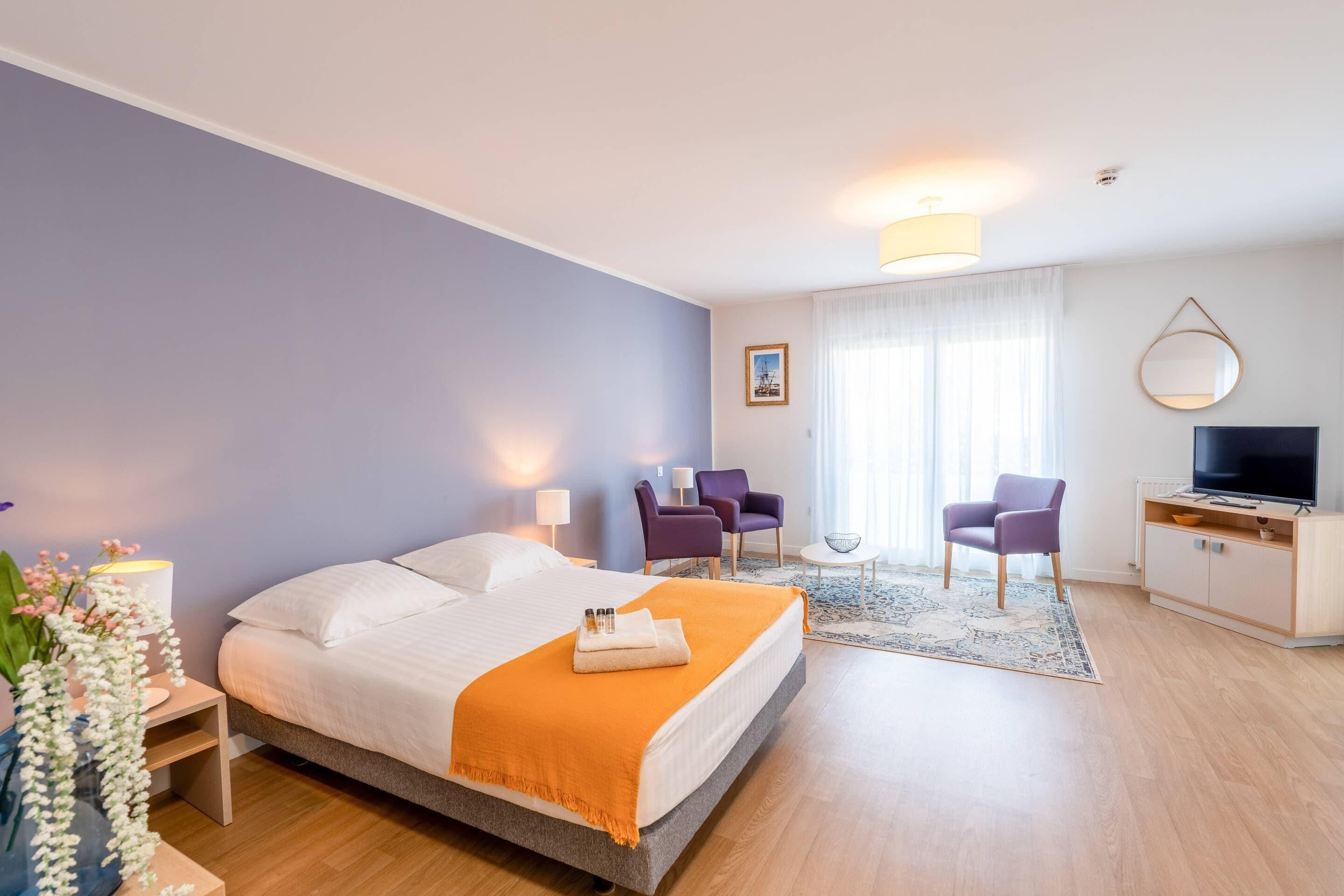 Ein modernes Hotelzimmer mit einem großen Bett, einem Schreibtisch mit Fernseher, zwei violetten Sesseln und einem kleinen Tisch mit einer Schale.