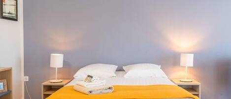 A neatly made bed with white linens and a yellow throw blanket, flanked by two bedside tables with lamps, in a room with a light blue wall.