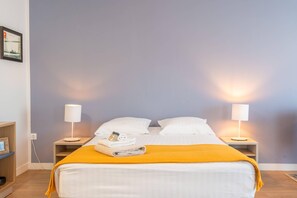 A neatly made bed with white linens and a yellow throw blanket, flanked by two bedside tables with lamps, in a room with a light blue wall.