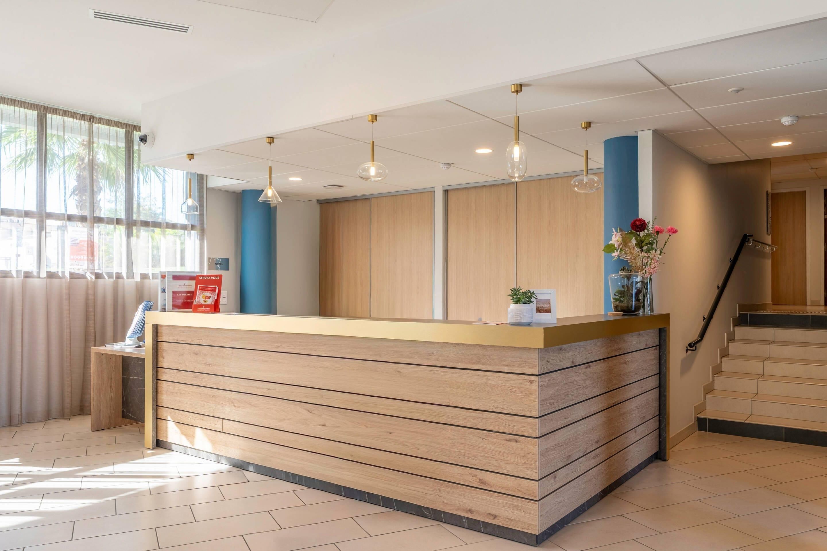 Un espace d’accueil doté d’un comptoir de réception en bois, d’un éclairage suspendu, d’un escalier et offrant une vue sur l’extérieur.