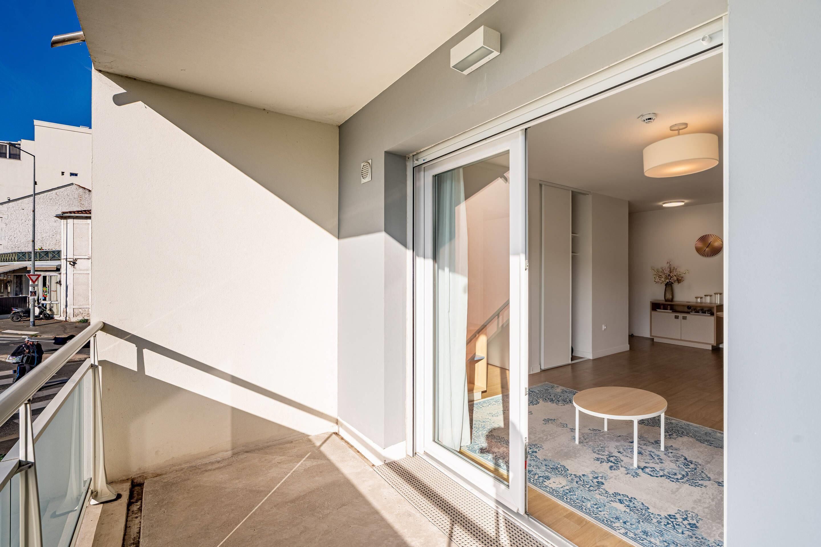 Un apartamento moderno con balcón, una puerta corredera de vidrio que da a una sala de estar y una cocina visible a través de la puerta.