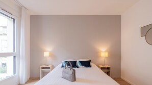 A hotel room with a large bed, two bedside tables with lamps, a window with curtains, and a handbag on the bed. - Quality Aparthotel Bourgoin-Jallieu (Bourgoin-Jallieu)