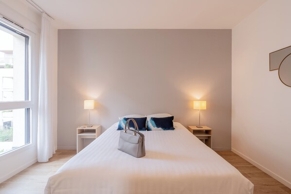 Ein Hotelzimmer mit einem großen Bett, zwei Nachttischen mit Lampen, einem Fenster mit Vorhängen und einer Handtasche auf dem Bett.
