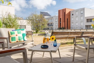 Un área de estar al aire libre con una mesa, sillas y un banco. Se observan edificios en el fondo, uno de ellos con un panel solar.