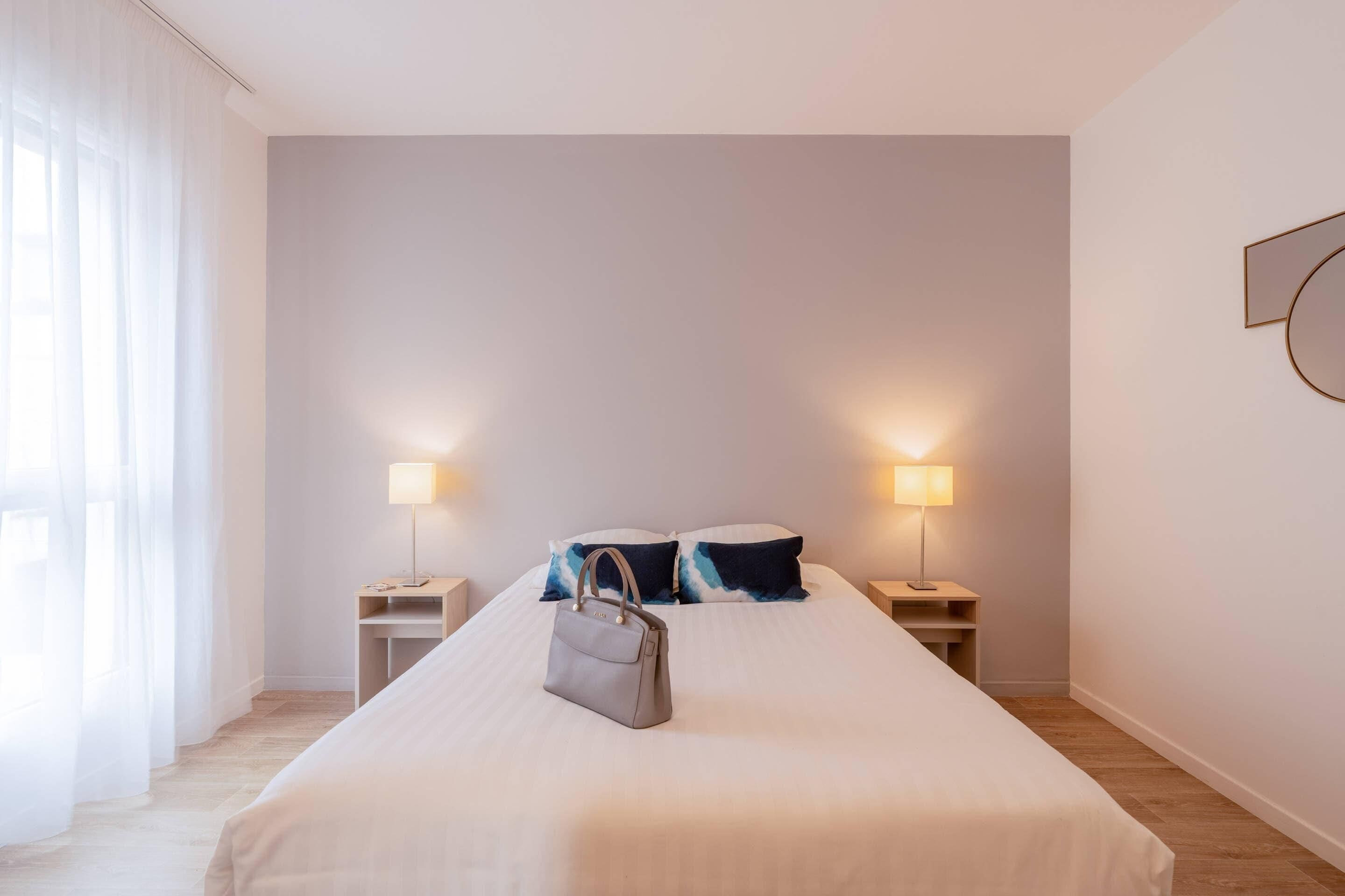 Habitación de hotel con una cama grande, dos mesitas de noche con lámparas, un bolso sobre la cama y cortinas translúcidas en la ventana.