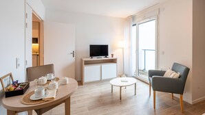 Ein modernes Wohnzimmer mit einem Essplatz, gedeckt für Tee, einem Fernseher auf einer Kommode, einem Sofa und einem großen Fenster mit Blick.