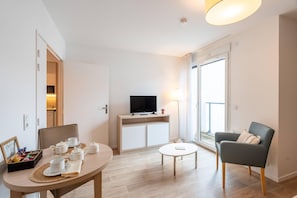 Un salon moderne avec une table à manger dressée pour le thé, un téléviseur posé sur un meuble, un canapé et une grande fenêtre offrant une vue dégagée.