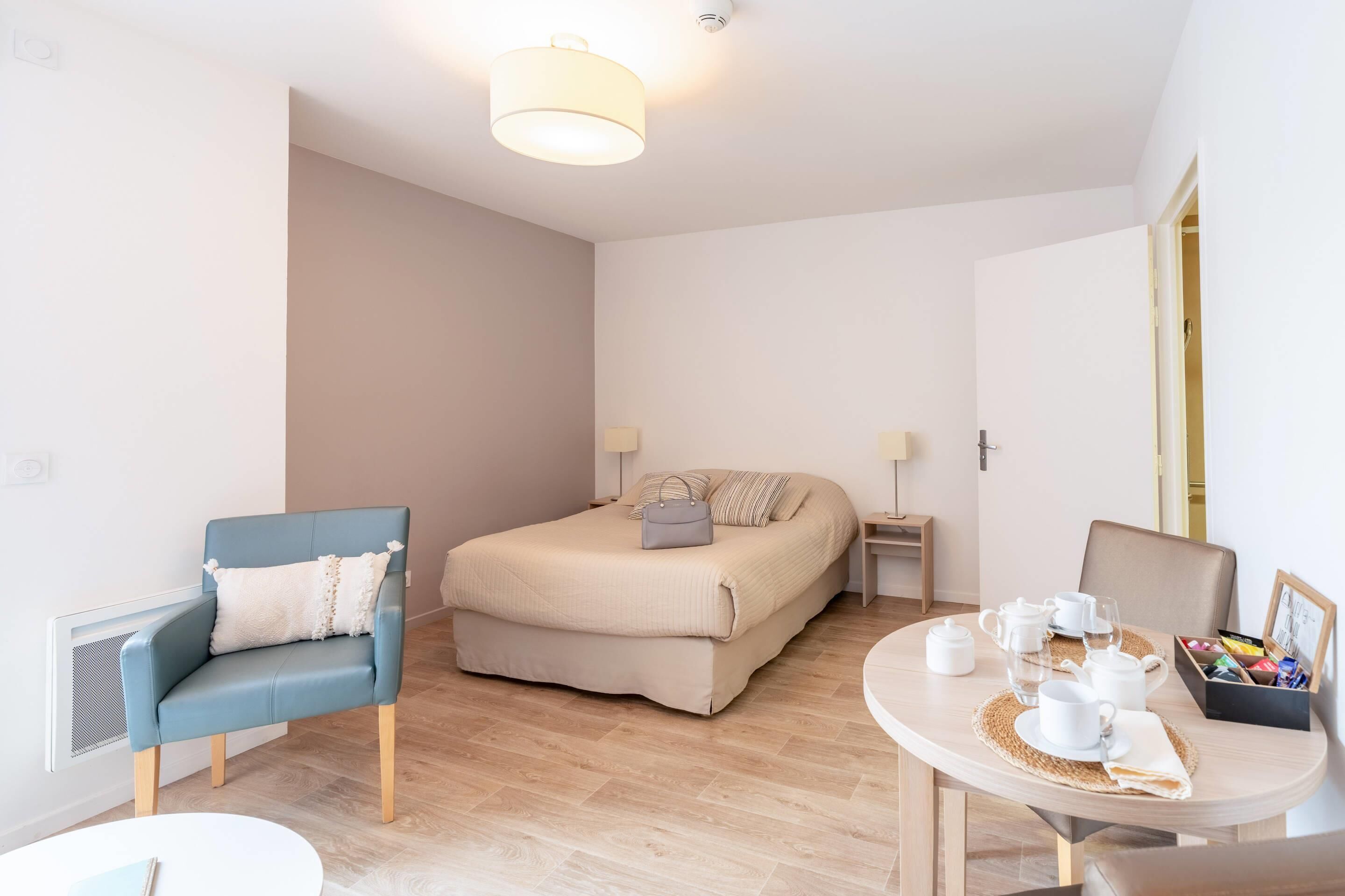 Una habitación moderna y luminosa con una cama, un sillón azul, una mesa redonda con juego de té y una mesita auxiliar.