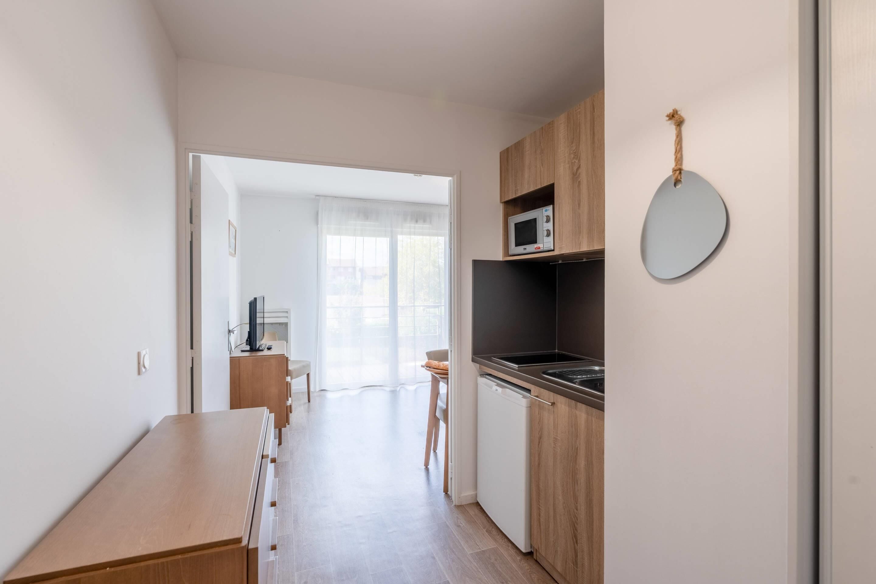 A compact kitchen with a microwave, sink, and wooden cabinets. A dining table and chair are visible in the adjacent room.
