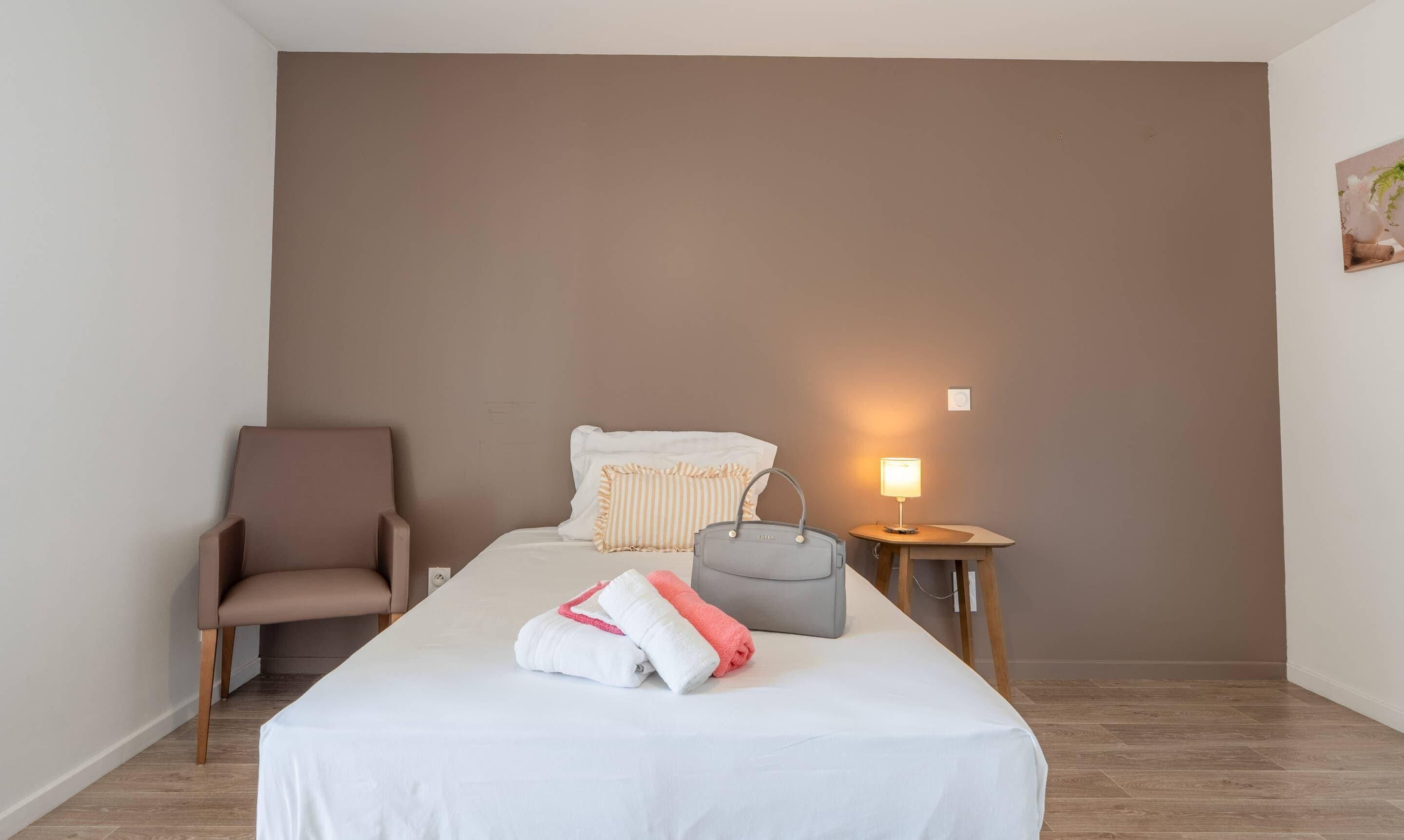 A hotel room with a bed, a chair, a bedside table with a lamp, and a wall-mounted plant.