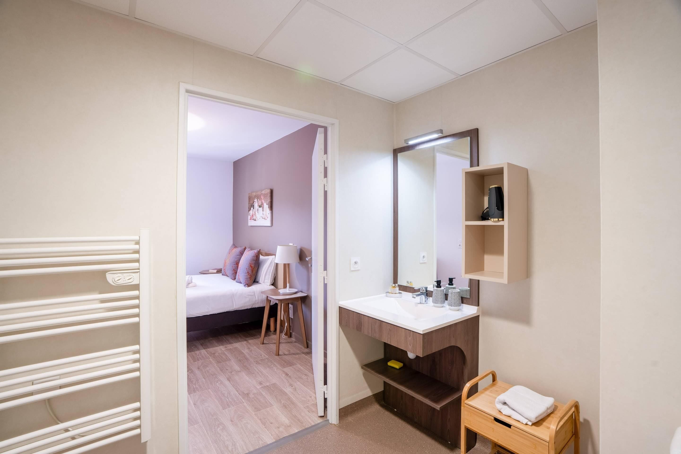 Hotellihuone, jossa on sänky, yöpöytä, lamppu sekä kylpyhuone, jossa on pesuallas, peili ja pyyheteline.