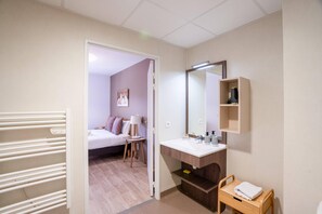 A hotel room with a bed, bedside table, lamp, and a bathroom with a sink, mirror, and towel rack. - Quality Aparthotel Lyon L'Isle-d’Abeau (L'Isle-d'Abeau)