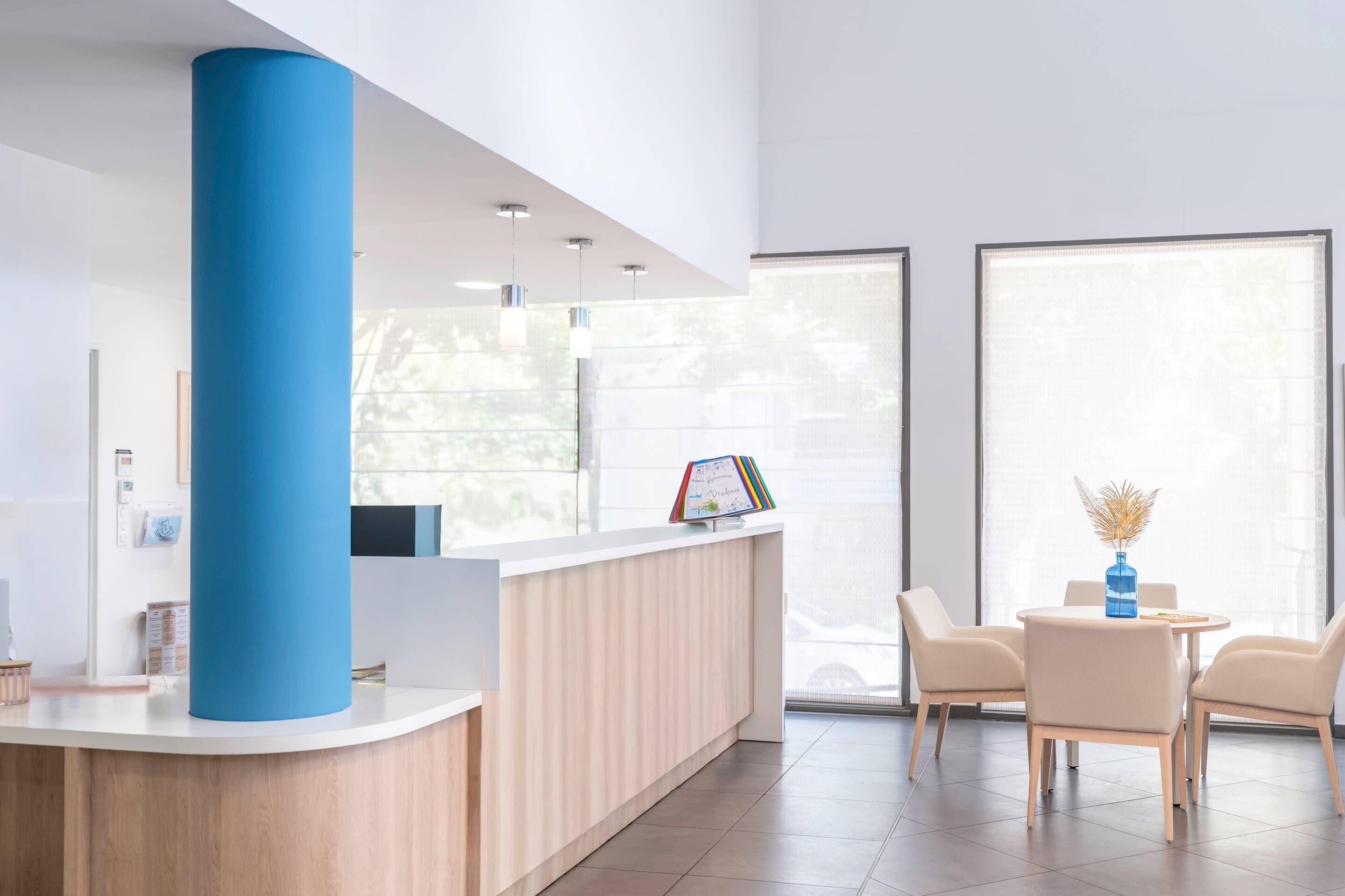 A modern reception area with a wooden reception desk, a blue accent column, and a seating area with two chairs and a small table.