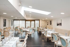 Un vaste espace dédié à la salle à manger, avec de nombreuses tables et chaises, de grandes fenêtres dotées de rideaux et des œuvres d’art accrochées aux murs.
