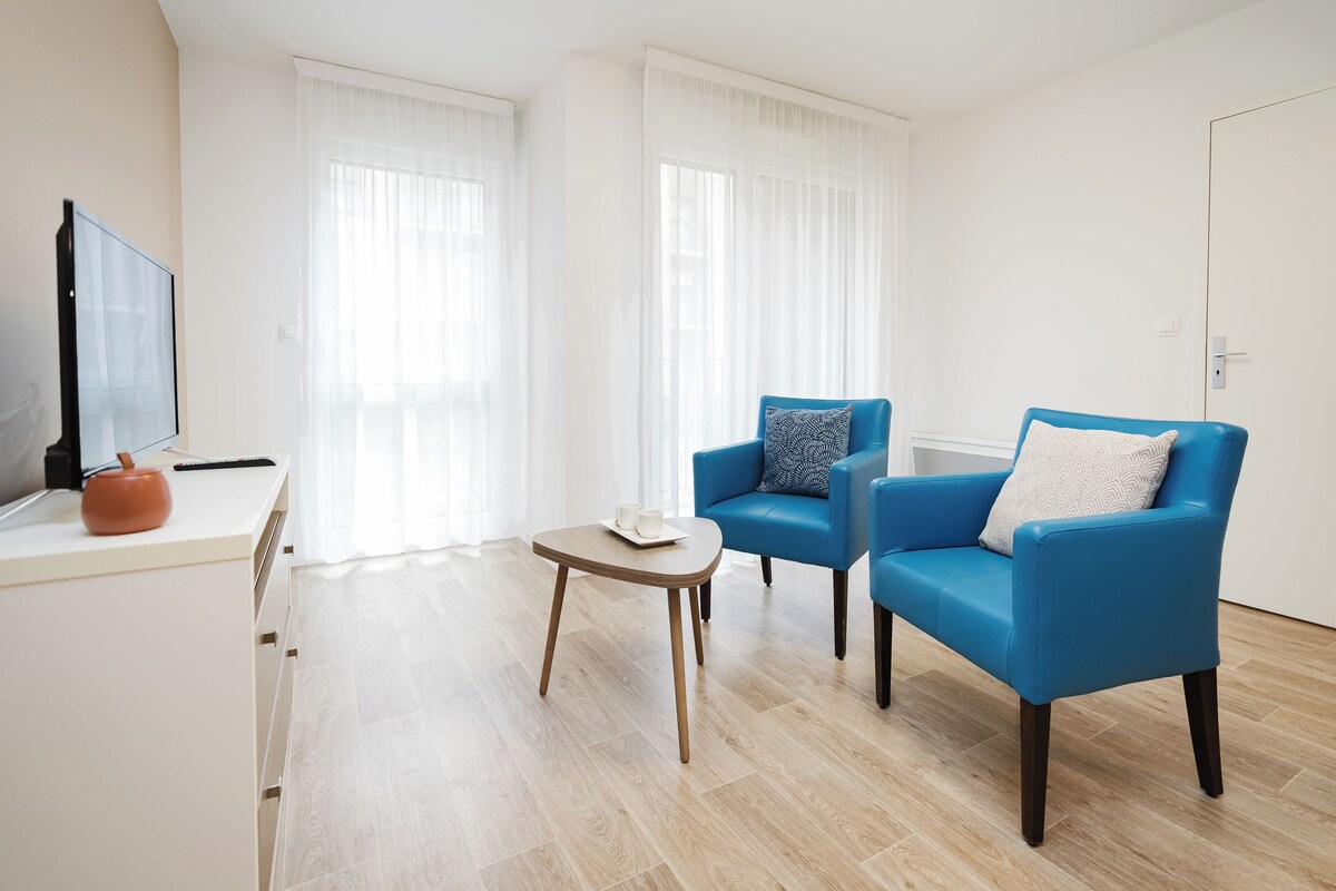 Een moderne woonkamer met een flatscreen televisie, twee blauwe fauteuils, een klein houten tafeltje met een dienblad en een witte kast.