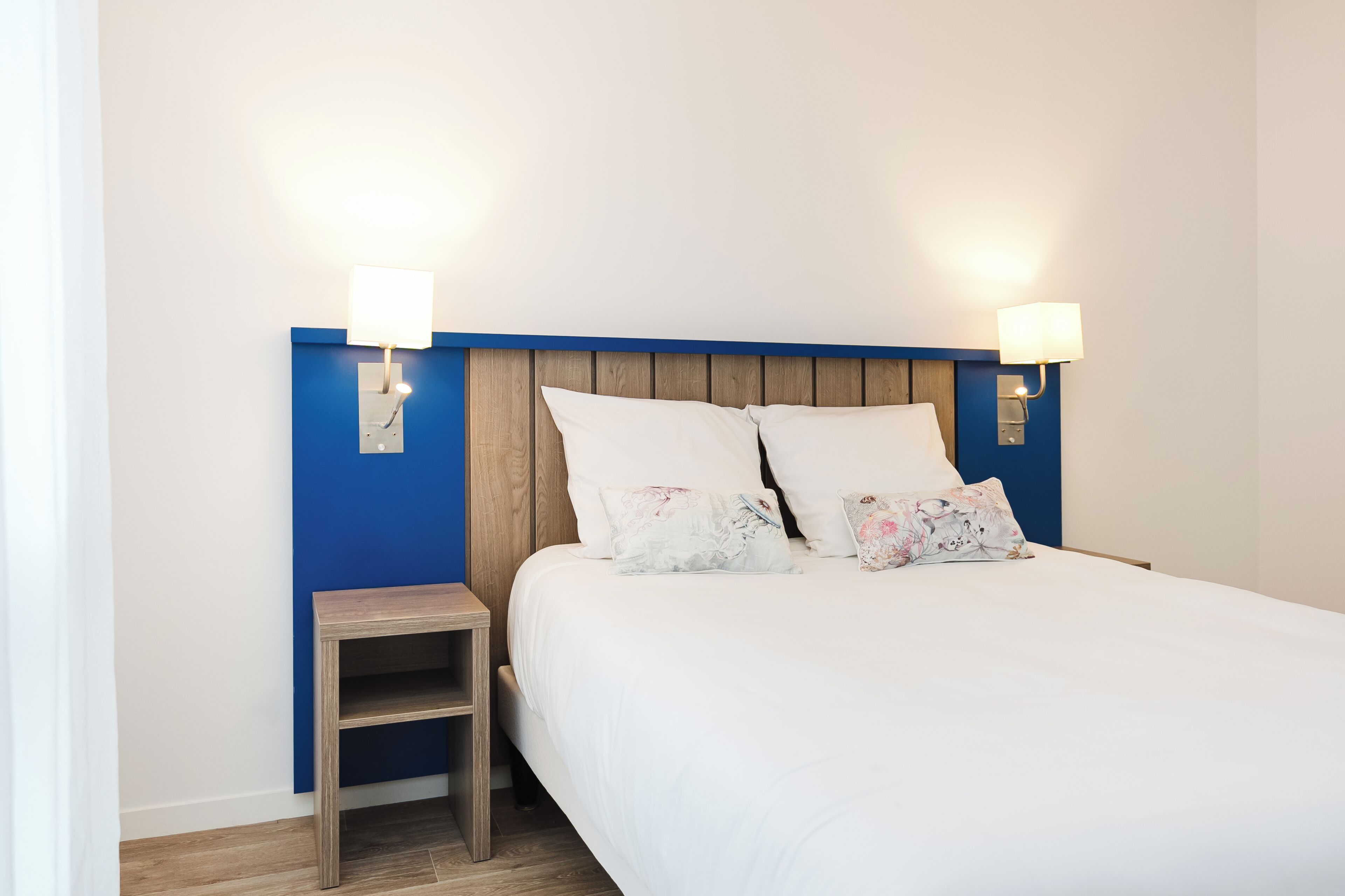 A neatly made bed with white linens and decorative pillows, a wooden nightstand, and wall-mounted lamps.
