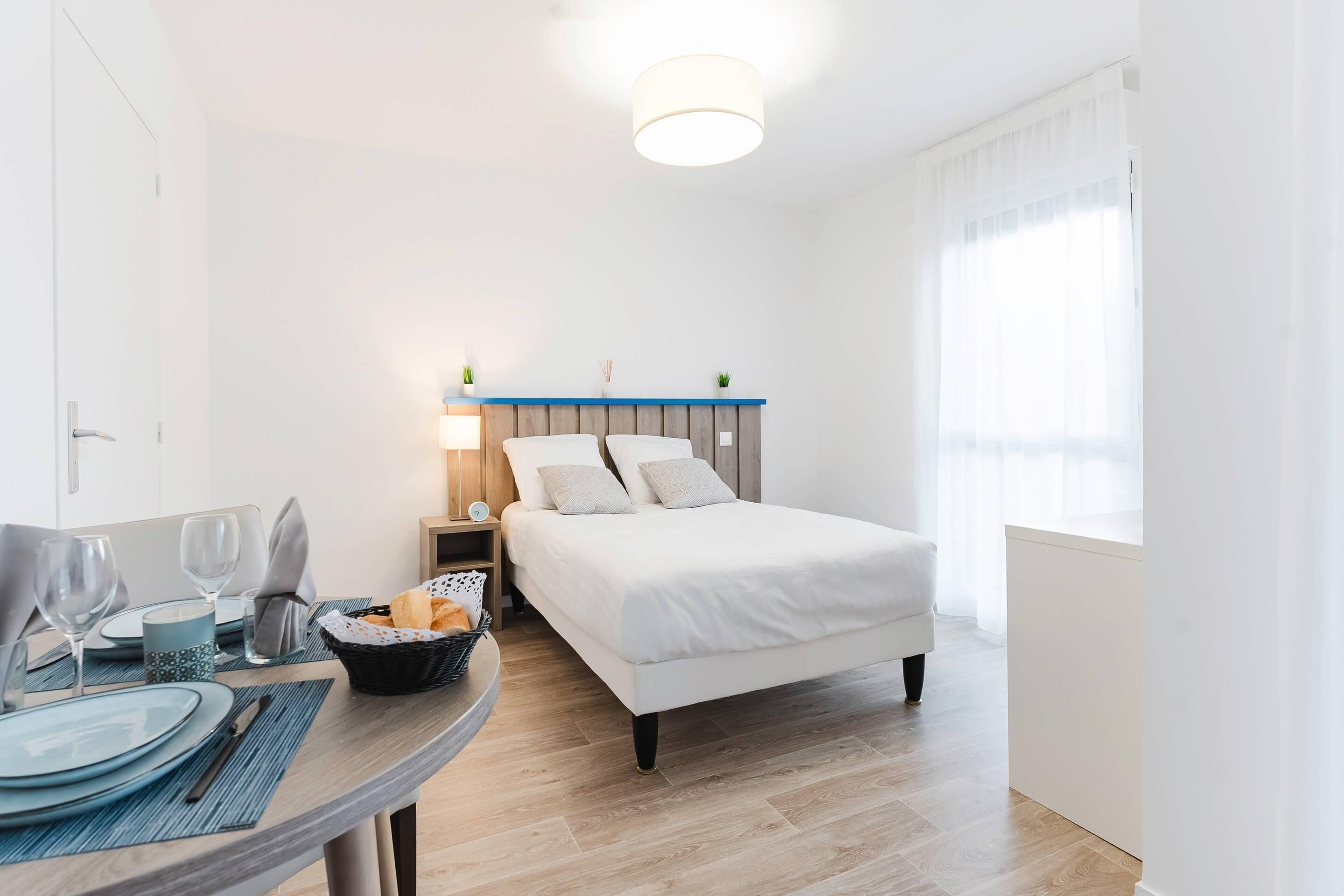 Un dormitorio ordenado con cama, mesita de noche, lámpara y un área de comedor con mesa puesta para dos.