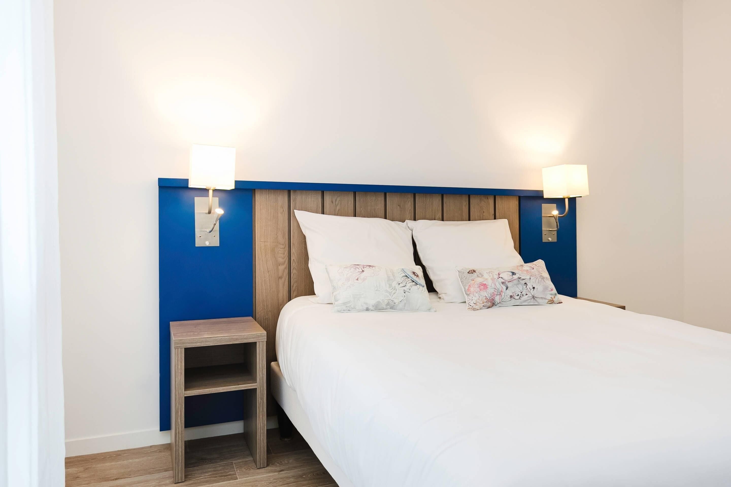 A neatly made bed with white bedding and two decorative pillows, a small wooden nightstand beside it, and two wall-mounted lamps on either side of the bed.