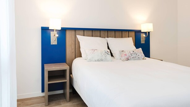 Een netjes opgemaakt bed met witte beddengoed en twee decoratieve kussens, een klein houten nachtkastje ernaast, en twee wandlampen aan weerszijden van het bed.