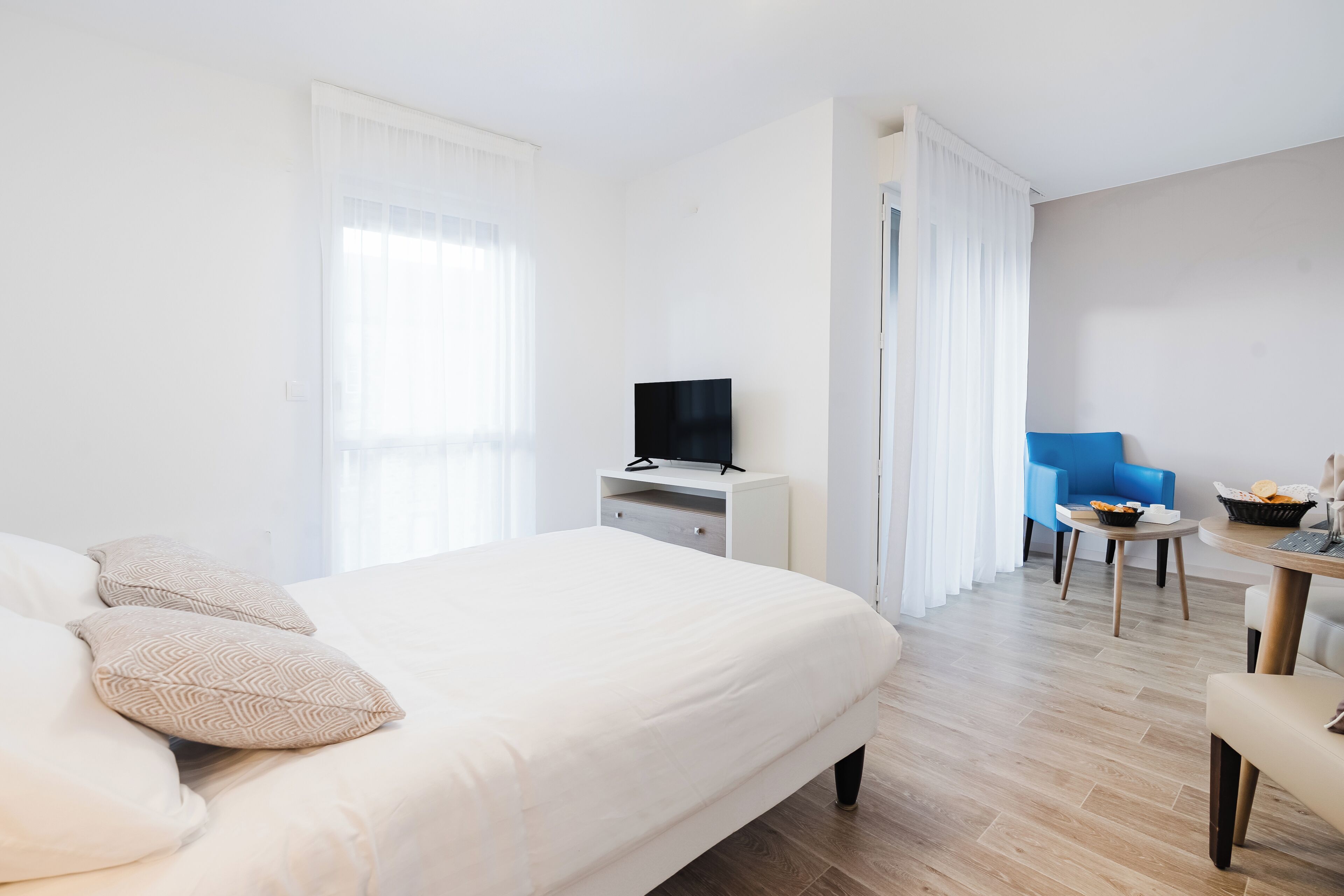 A hotel room with a bed, a desk with a TV, a chair, and a small table with snacks.
