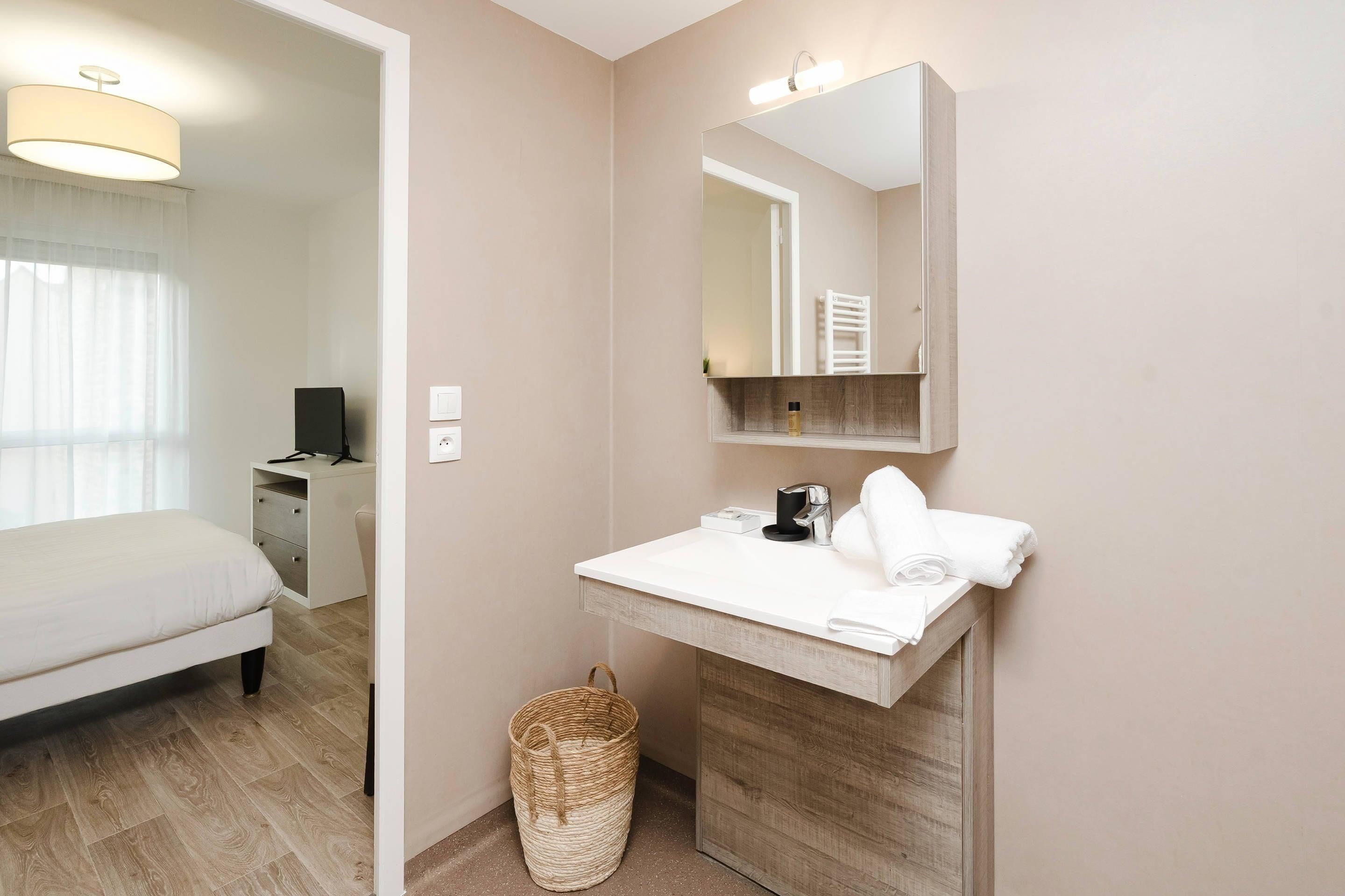Une chambre d’hôtel moderne avec un lit, un lavabo blanc, un miroir et une armoire en bois.