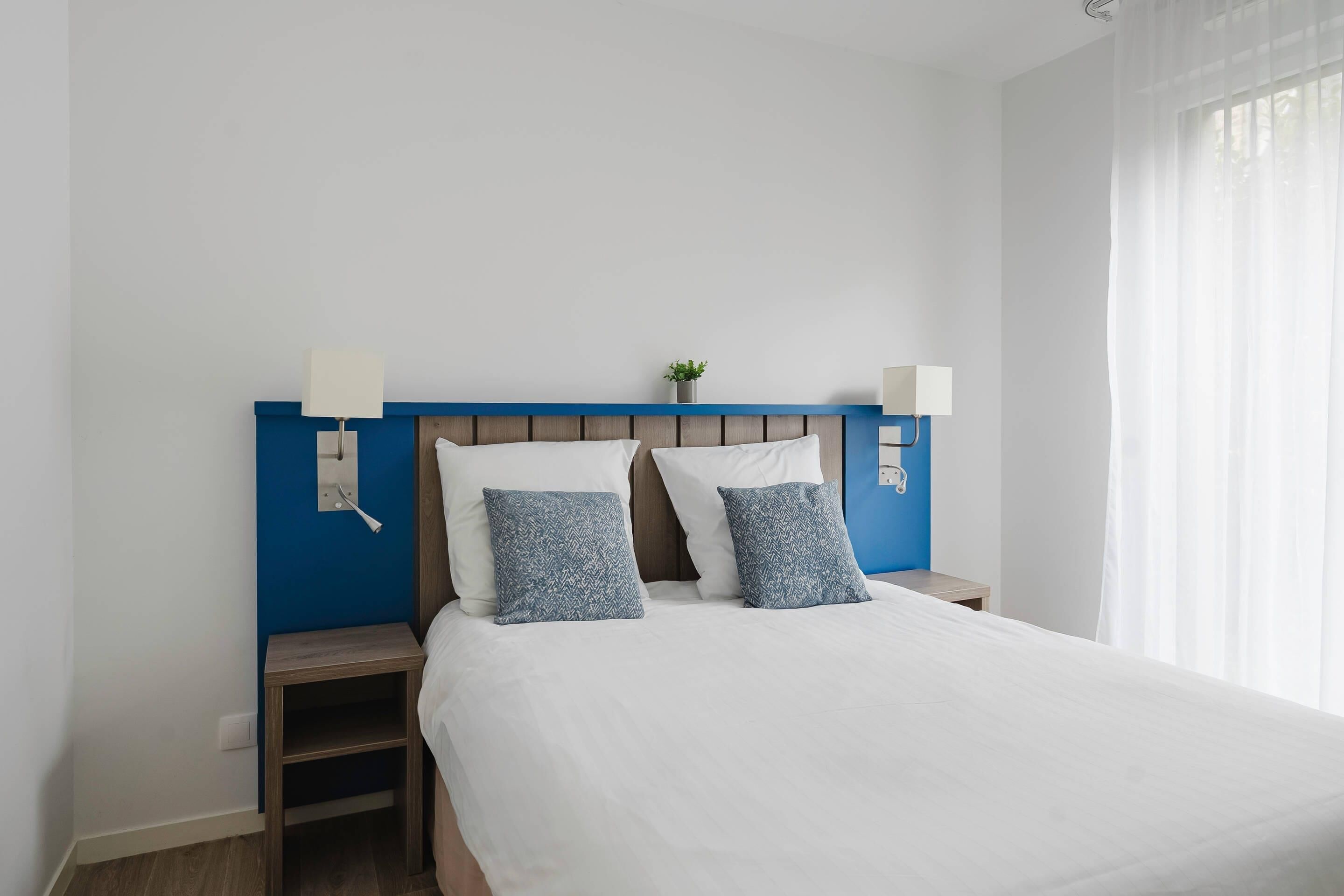 Un lit bien fait, avec une literie blanche et deux coussins à motifs bleus, une tête de lit en bois et deux tables de chevet avec des lampes.
