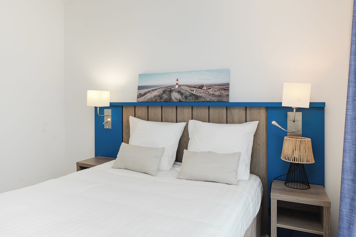 Una cama bien hecha con sábanas blancas, cabecera con panel azul y un cuadro colgado en la pared de un faro.